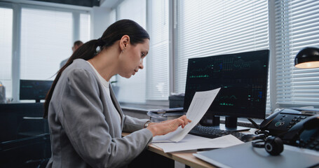 Beautiful adult female financial specialist working with papers, searching and analyzing data, graphs and charts of financial metrics changing on computer monitor. Working in modern business office.