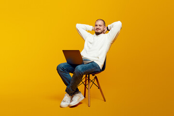 Full body of happy man sitting on chair using laptop computer, holding hands behind neck, rest, relax, isolated over yellow background