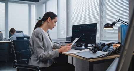 Fototapeta premium Workplace of managers of large financial company: Adult, beautiful female analyst checking data in paper documents for changes in foreign exchange rates. Monitoring stock and equity markets concept.