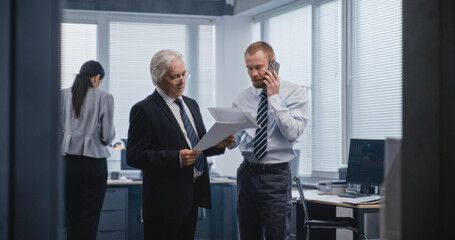 Serious professional stock trader, stockbroker speaks with business client on call using mobile phone. Senior boss of the company stands near and listens to conversation. Work in modern office.