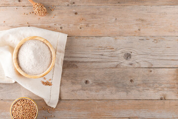 Buckwheat flour in a wooden bowl and raw buckwheat grain on cotton napkin on rustic wooden table background, top view, copy space. Alternative flour, gluten free flour, healthy nutrition