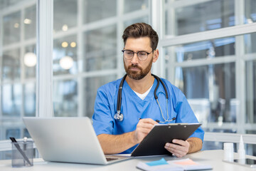 Focused doctor working with laptop and clipboard in modern office. Wearing blue scrubs, glasses, and stethoscope. Concept of healthcare, professionalism, medical consultation, technology in medicine.