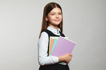 Portrait of happy positive pupil girl in school uniform carrying textbook and looking at camera against white background. Back to school concept