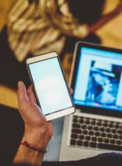 Cropped image of male using laptop computer and netbook for working online via 4G connection, man holding smartphone with mock up screen confirming payment via transaction on mobile phone app