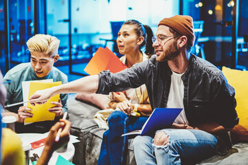 Crew of positive male and female employees working in team cooperating and share ideas, hipster guy pointing on smth while brainstorming on meeting with students group in modern coworking space