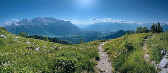 A winding dirt path leads through lush green hills with a view of majestic mountains and a valley in the distance under a clear blue sky.