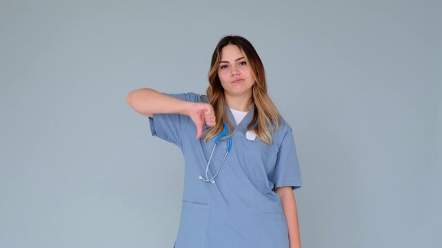 Dissatisfied and serious female Caucasian doctor or nurse showing thumb down gesture in rejection, decline, standing on gray background.