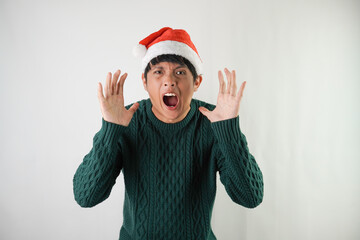 Angry young asian man wearing santa clause hat and green long sleeved sweater, isolated over white background. Concept for Christmas Holiday and New Year Party