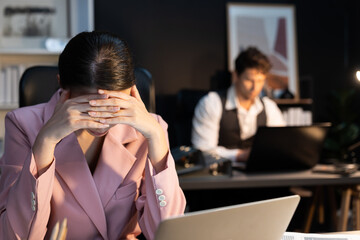 Smart businesswoman having aches with body in back, neck and whole body, stretching composition in long time with man working at blurry back site at neon dark room at late overnight time. Postulate.