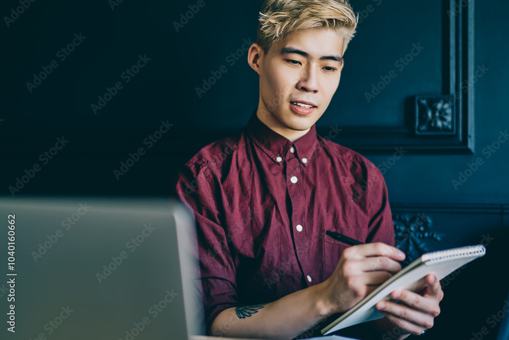 Poster pensive blonde japanese hipster guy holding notepad in hand and writing down checklist sitting at la