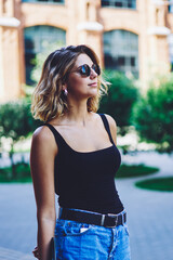 Positive female traveller in fashionable sunglasses enjoying journey during summer holidays, attractive stylish woman spending free time in college campus, concept of recreating and vacations