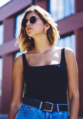 Serious attractive female student in stylish sunglasses looking away and thinking about free time during sunny summer day, beautiful female teenager casual dressed resting after walking in city