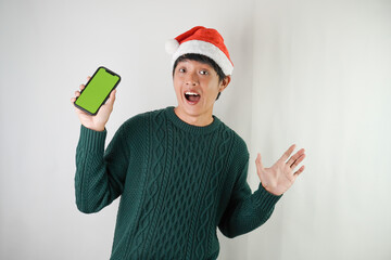 Excited young asian man wearing santa clause hat and green long sleeved sweater is showing smartphone green screen, isolated over white background. Concept for Christmas Holiday and New Year Party
