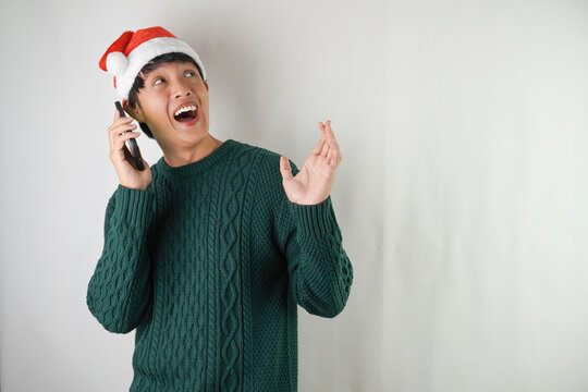 Excited young asian man wearing santa clause hat and green long sleeved sweater is calling someone using smartphone, isolated over white background. Concept for Christmas Holiday and New Year Party