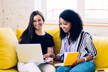 Cheerful multiracial women sitting with laptop on yellow sofa