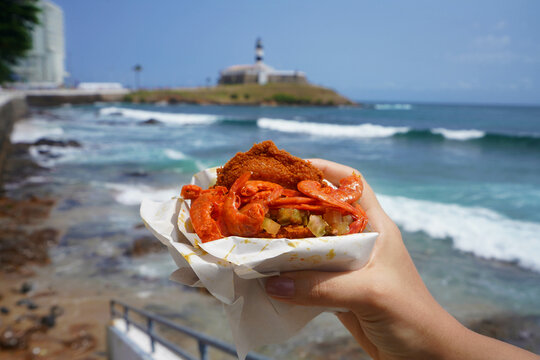 Acaraje traditional dish and street food from Northeast Brazil with Farol da Barra Lighthouse and Bay of All Saints on the background, Salvador de Bahia, Brazil