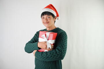 Excited young asian man wearing santa clause hat and green long sleeved sweater is carrying large gift box with ribbon, isolated over white background. Concept for Christmas Holiday and New Year Party