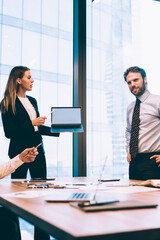 Intelligent female employee in elegant suit holding modern laptop computer with mock up area for advertising text for financial website while having brainstorming process with professional colleagues