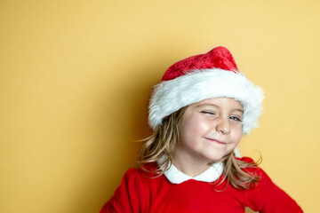 Little girl with funny gesture winks dressed as Santa Claus on yellow background.