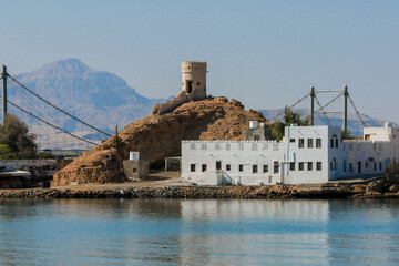 A coastal view of Sur Oman showcasing a historic tower and a bridge over calm waters during midday