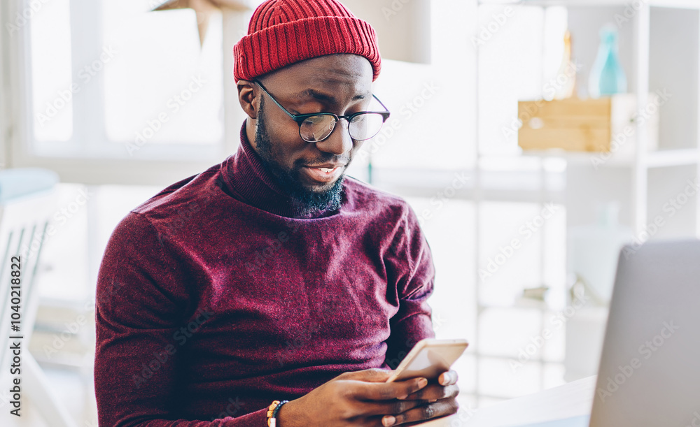 Poster african american hipster guy in spectacles sending text message on mobile phone while working online