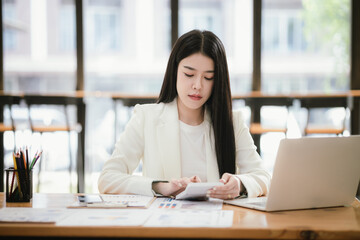 Beautiful Asian female accountant working with smartphone and laptop at her desk analyzing business reports and documents. Sending messages