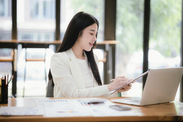 Beautiful Asian female accountant working with smartphone and laptop at her desk analyzing business reports and documents. Sending messages