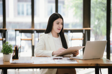 Beautiful Asian female accountant working with smartphone and laptop at her desk analyzing business reports and documents. Sending messages