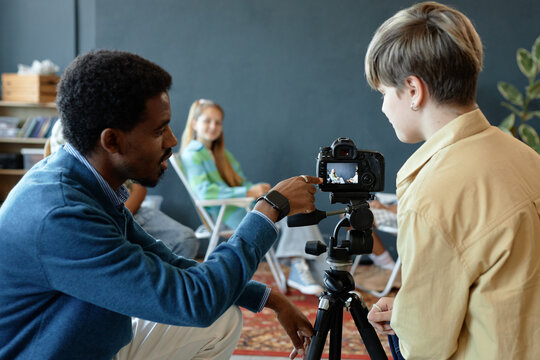 Back view of teenage boy setting up camera on tripod in studio with teacher or mentor explaining settings and pointing at digital screen
