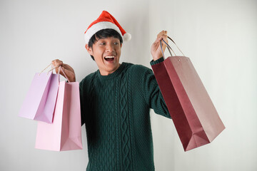 Excited young asian man wearing santa clause hat is carrying many shopping bag, isolated over white background. Concept for Christmas Holiday and New Year Party.