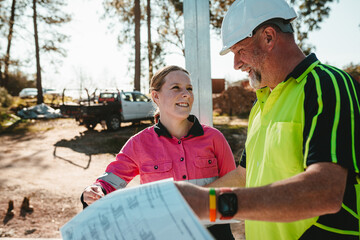 Apprentice builder laughing with  tradesman while looking at the blueprint on worksite