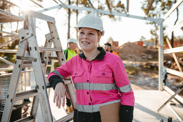 A young woman standing on worksite resting arm on ladder wearing hard hat