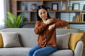 Asian woman experiencing shoulder pain while sitting on sofa in home interior. She touches sore...