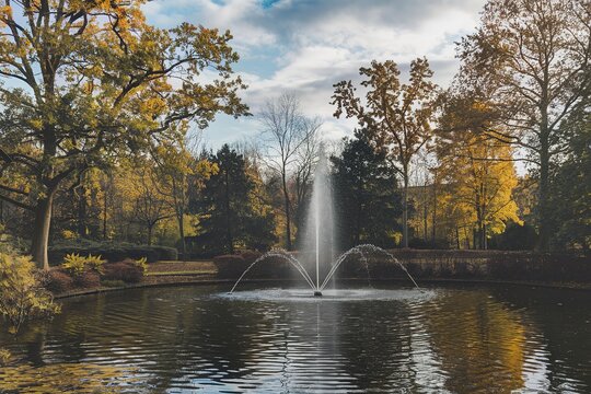 A scenic park with a large fountain in the center of a pond surrounded by trees with autumn foliage