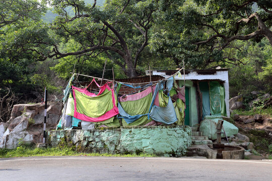 A small, rustic structure draped with colorful fabrics, nestled in a lush, green forest with a hand pump by the entrance.