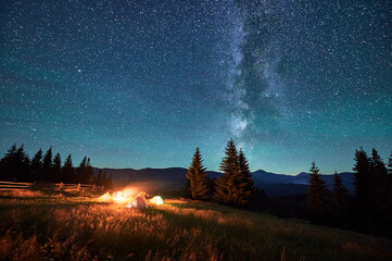 Silhouettes of tourists resting at campsite around campfire with glowing tents nearby under starry...