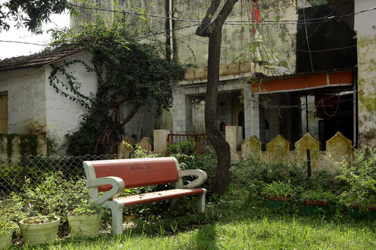 A quiet, charming spot with a red bench amidst lush greenery, set beside an old, vine-covered house. The scene exudes a sense of calm and nature&rsquo;s embrace.