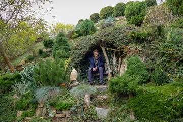 boy sits in a decorative gazebo on a green slope, bushes, stones, plants, landscape
