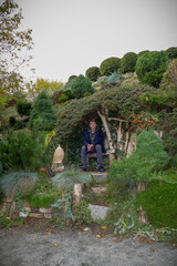 boy sits in a decorative gazebo on a green slope, bushes, stones, plants, landscape