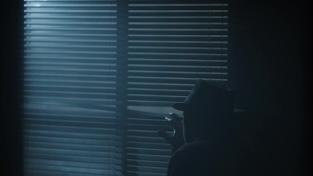 Silhouette of a man dressed in long coat with hat walking to a dark room window. High quality 4k footage