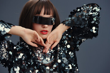 A woman in a sequinned top and sunglasses with her hands on her face looks directly at the camera,...