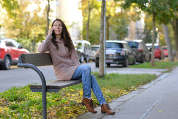 A woman talking on a phone sitting on a bench at the street on a sunny autumn day. A woman smiles while using a mobile smartphone device