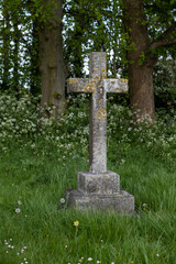 An old weathered and moss covered cross framed by two trees in old churchyard, surrounded by grass and wild flowers