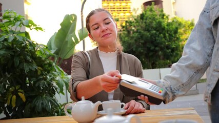 Smiling young woman sitting at a bar terrace, paying for her tea using a credit card and contactless payment terminal, highlighting convenience and modern technology in outdoor dining settings