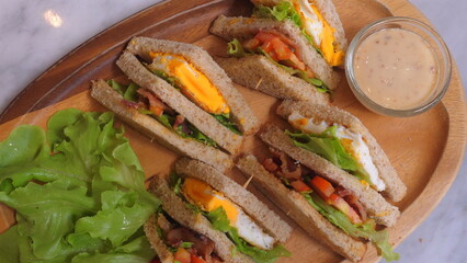 Delicious club sandwiches with eggs, bacon, lettuce, and tomato on whole wheat bread served on wooden platter, accompanied by fresh green lettuce and small dish of creamy dressing. Food and Cuisine.
