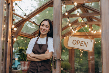 Happy floral shop owner standing with arms crossed on chest at doorway of empty glasshouse full of plants, local small business concept
