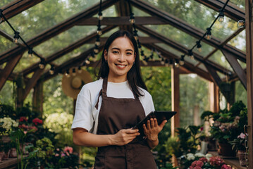 Portrait of smiling dark haired lady standing at greenhouse and holding digital tablet in hands
