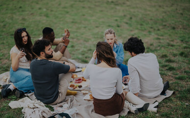 A group of friends gathered for a picnic, sitting on a blanket in a lush green field. They are enjoying food, drinks, and each other's company on a pleasant day.