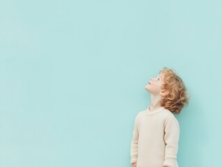 A small child isolated on a light blue background. A red-haired boy in white clothes against a blue wall looks sideways and upward. Child in profile looking up, copy space.