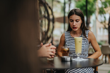 A young woman in a striped dress sits at an outdoor cafe table with drinks, appearing relaxed and engaged in conversation with a friend.